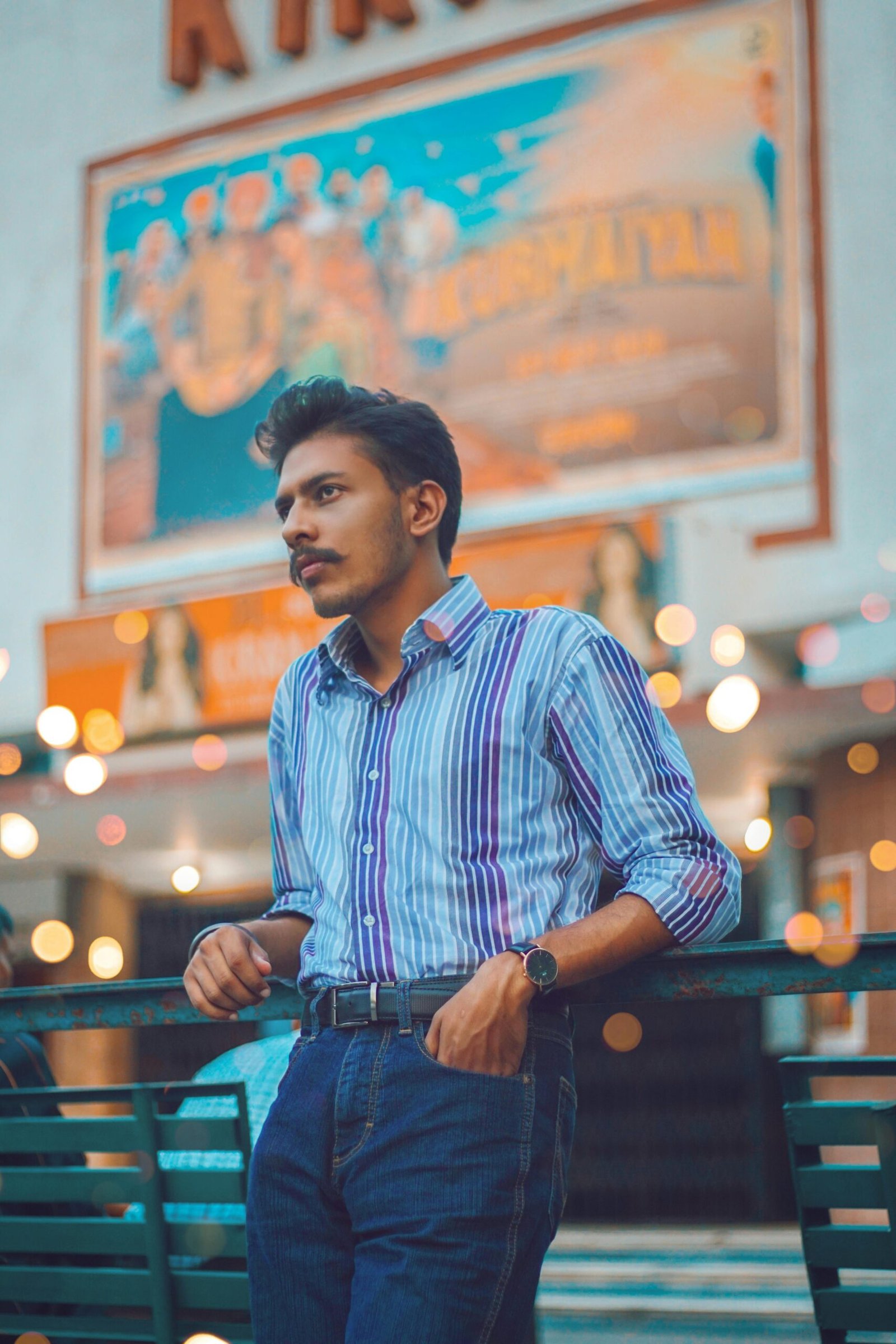 Young man with moustache in striped shirt posing at night with bokeh lights background.